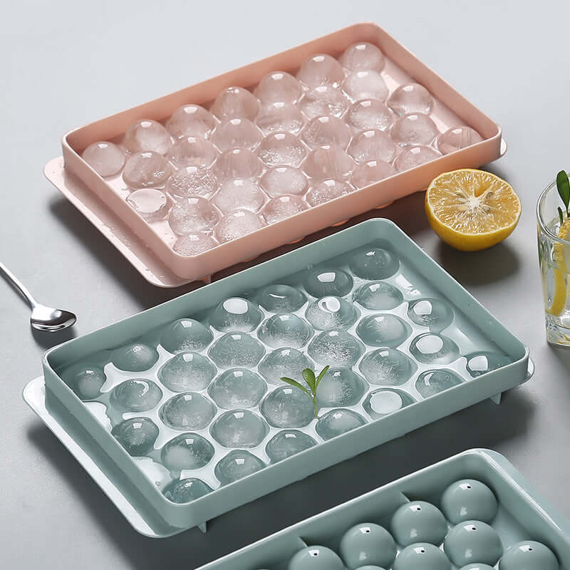 3D round ice cube mold in pink and blue trays with ice, lemon, and a spoon on a grey background.